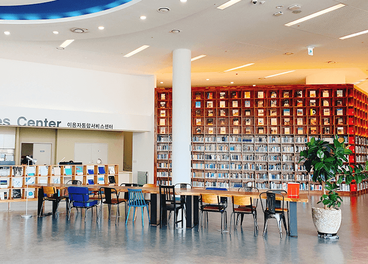 해양도서관 내부 전경, 중앙 긴 나무 테이블에 다양한 색상의 의자들이 배치되어 있고, 사진 왼편에 이용자 통합서비스센터가 오른쪽에는 각종 도서들이 진열되어 있는 모습.