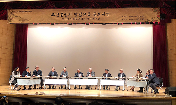 강당 위에 테이플을 놓고 의자에 앉아서 앞을 바라보며 회의하고 있는 사람들의 모습
